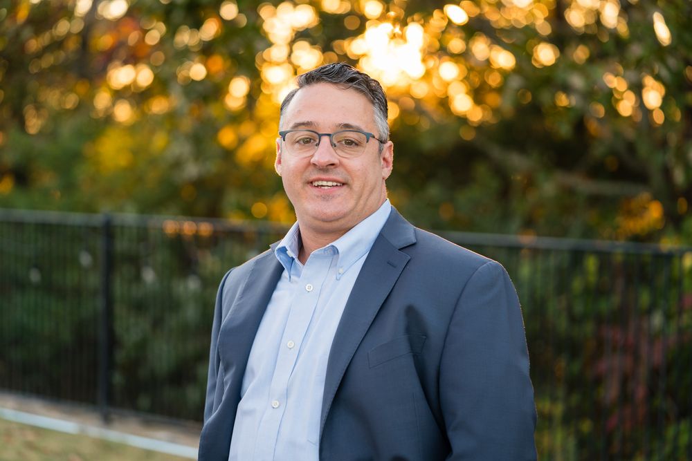 A man in a blue suit and glasses stands outdoors, smiling, with a blurred fence and trees in the background at sunset.