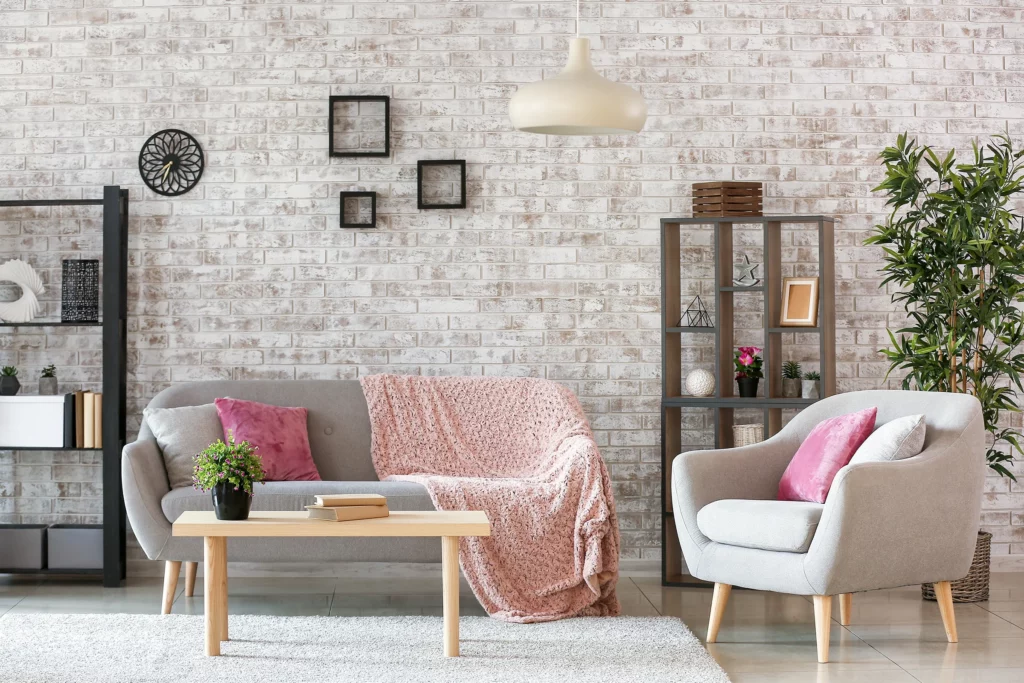 Modern living room with a gray sofa, armchair, pink accents, wooden coffee table, shelves, potted plants, and a white brick wall—perfect inspiration for budget-friendly tips to stage or sell your home.