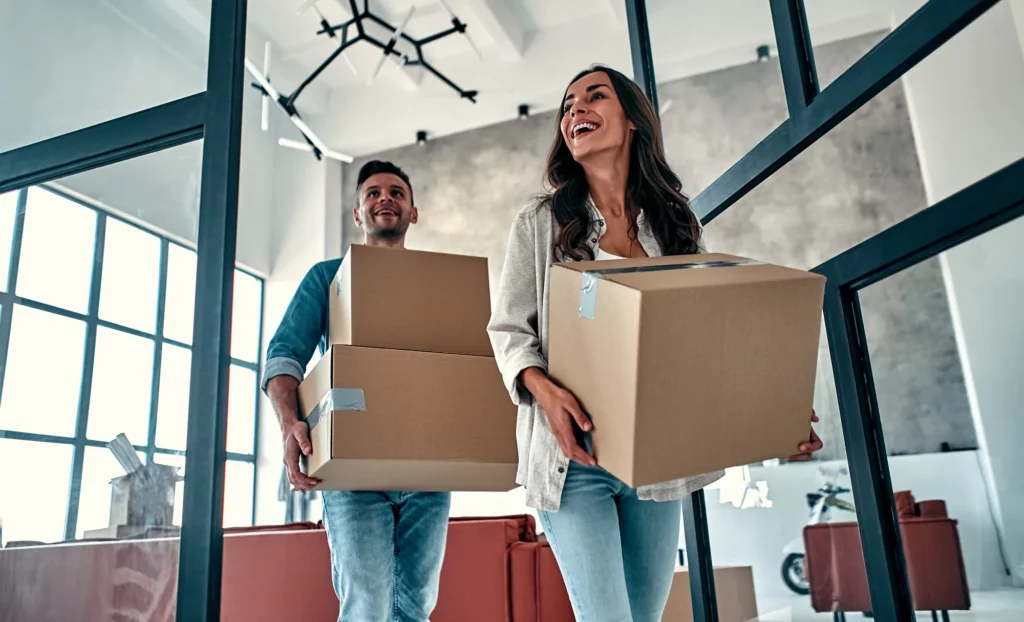 Two people smiling and carrying cardboard boxes walk into a modern, well-lit room with large windows—a perfect scene for Central Arkansas real estate and happy home buyers starting a new chapter.