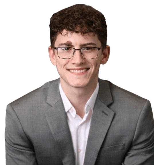 Heath Besowshek, a young man with curly brown hair and glasses, smiles at the camera in a gray suit jacket and white shirt against a plain background.